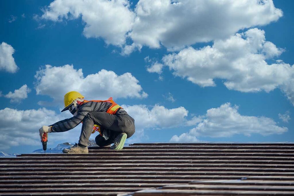 A worker installs new asphalt shingles on a pitched house roof under partly cloudy skies.